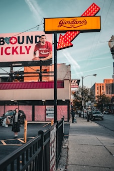 A street scene captures a Giordano's sign with bright red and yellow colors, featuring an illuminated arrow. A large advertisement billboard is visible on the left, displaying a man in a red shirt holding a basketball, partially obscured by the sign. Below the billboard, a woman stands in a parking lot holding shopping bags with a smile, dressed in dark clothing. On the right side of the image, a person with a suitcase walks down the sidewalk beside a street with visible cars. A 'For Lease' sign is posted on the building in the background.