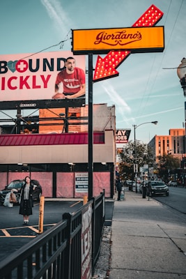 A street scene captures a Giordano's sign with bright red and yellow colors, featuring an illuminated arrow. A large advertisement billboard is visible on the left, displaying a man in a red shirt holding a basketball, partially obscured by the sign. Below the billboard, a woman stands in a parking lot holding shopping bags with a smile, dressed in dark clothing. On the right side of the image, a person with a suitcase walks down the sidewalk beside a street with visible cars. A 'For Lease' sign is posted on the building in the background.