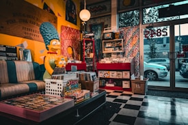 A cozy, eclectic record store with a retro vibe. A large statue of a blue-haired cartoon character is prominently displayed, sitting on a vintage-style sofa. The shelves are filled with vinyl records and colorful posters, enhancing the nostalgic atmosphere. Bright, multicolored patterns adorn the walls, and a large window shows a view of parked cars outside.