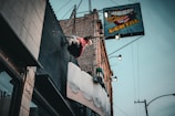 A dental office sign with a large toothbrush and oversized teeth protrudes from the side of a brick building. Above the sign, a mannequin styled like a trapeze artist swings from a trapeze bar, adding a whimsical element to the scene. The sky is overcast, and the building facade features various textures such as metal and stone.