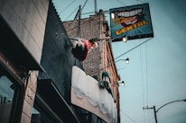A dental office sign with a large toothbrush and oversized teeth protrudes from the side of a brick building. Above the sign, a mannequin styled like a trapeze artist swings from a trapeze bar, adding a whimsical element to the scene. The sky is overcast, and the building facade features various textures such as metal and stone.