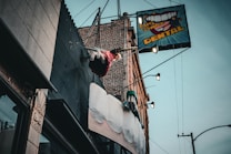 A dental office sign with a large toothbrush and oversized teeth protrudes from the side of a brick building. Above the sign, a mannequin styled like a trapeze artist swings from a trapeze bar, adding a whimsical element to the scene. The sky is overcast, and the building facade features various textures such as metal and stone.