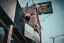 A dental office sign with a large toothbrush and oversized teeth protrudes from the side of a brick building. Above the sign, a mannequin styled like a trapeze artist swings from a trapeze bar, adding a whimsical element to the scene. The sky is overcast, and the building facade features various textures such as metal and stone.