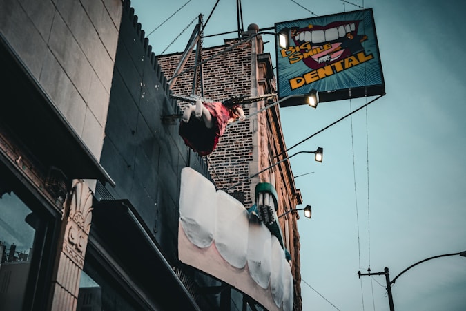 A dental office sign with a large toothbrush and oversized teeth protrudes from the side of a brick building. Above the sign, a mannequin styled like a trapeze artist swings from a trapeze bar, adding a whimsical element to the scene. The sky is overcast, and the building facade features various textures such as metal and stone.