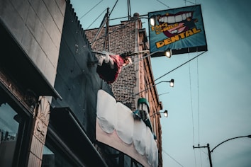 A dental office sign with a large toothbrush and oversized teeth protrudes from the side of a brick building. Above the sign, a mannequin styled like a trapeze artist swings from a trapeze bar, adding a whimsical element to the scene. The sky is overcast, and the building facade features various textures such as metal and stone.