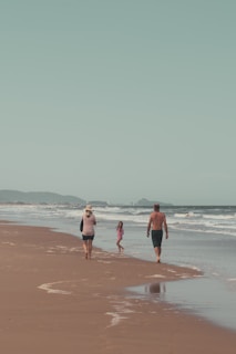 A family walks along a sandy beach near the shoreline. The ocean waves gently approach the shore and there are distant hills visible under a clear sky. The atmosphere is peaceful and relaxed, suggesting a leisurely day.