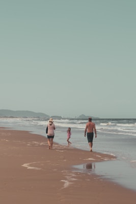 A family walks along a sandy beach near the shoreline. The ocean waves gently approach the shore and there are distant hills visible under a clear sky. The atmosphere is peaceful and relaxed, suggesting a leisurely day.