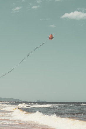 A colorful kite with a face design is soaring high in the sky above a beach. The ocean waves are gently crashing onto the shore, and the distant landscape includes some hills under a clear sky with a few clouds.