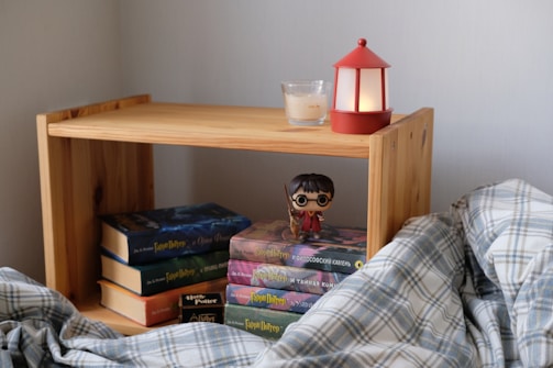 A small wooden bookshelf holds a stack of books, including several from the Harry Potter series. A figurine of a bespectacled character wearing a red robe is prominently placed on the books. A small red lantern and a lit candle sit on top of the shelf. A plaid blanket is partly visible in the foreground, adding a cozy element to the scene.