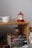 A wooden shelf holds several hardcover books stacked in a neat pile. On top of the books sits a small figurine wearing glasses and holding a broomstick. To the left on the shelf, there is a white ceramic mug and a clear glass containing a small amount of liquid. A red, decorative lantern with a white interior is lit beside them. The setting conveys a cozy and inviting atmosphere.