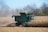 A large green combine harvester is actively working in a cornfield, cutting through dried corn stalks. Dust is stirred up around the machine, indicating the harvesting process. Bare trees surround the field, suggesting late autumn or early winter.