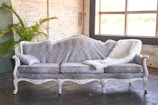 An elegant antique-style sofa upholstered in a light grey patterned fabric, adorned with a decorative pillow and a fluffy white throw. It is set against a white brick wall with a large window providing natural light. A green plant sits in the corner, adding a touch of nature.