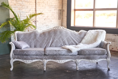 An elegant antique-style sofa upholstered in a light grey patterned fabric, adorned with a decorative pillow and a fluffy white throw. It is set against a white brick wall with a large window providing natural light. A green plant sits in the corner, adding a touch of nature.