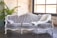 An elegant antique-style sofa upholstered in a light grey patterned fabric, adorned with a decorative pillow and a fluffy white throw. It is set against a white brick wall with a large window providing natural light. A green plant sits in the corner, adding a touch of nature.