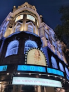 A night scene of the Novello Theatre with brightly lit signs and posters for the musical 'Mamma Mia!'. The historic architecture features tall columns and decorative windows. The main sign reads 'You Already Know You're Gonna Love It!' in bold letters.