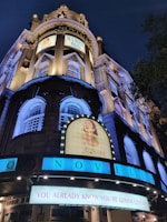A night scene of the Novello Theatre with brightly lit signs and posters for the musical 'Mamma Mia!'. The historic architecture features tall columns and decorative windows. The main sign reads 'You Already Know You're Gonna Love It!' in bold letters.