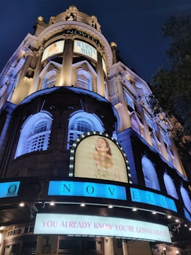 A night scene of the Novello Theatre with brightly lit signs and posters for the musical 'Mamma Mia!'. The historic architecture features tall columns and decorative windows. The main sign reads 'You Already Know You're Gonna Love It!' in bold letters.