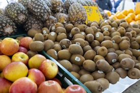 A market display featuring piles of fresh fruit. There are numerous kiwis in the center with small stickers on them. Apples with a mix of red and yellow hues are in the foreground. In the background, a stack of pineapples and some oranges are visible. The fruit is arranged neatly in containers, suggesting a vibrant and bustling market setting.