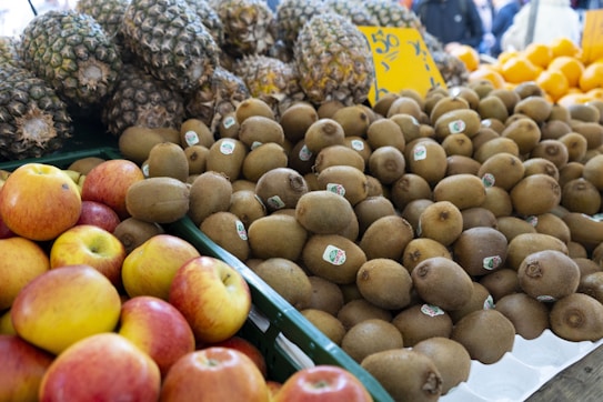 A market display featuring piles of fresh fruit. There are numerous kiwis in the center with small stickers on them. Apples with a mix of red and yellow hues are in the foreground. In the background, a stack of pineapples and some oranges are visible. The fruit is arranged neatly in containers, suggesting a vibrant and bustling market setting.