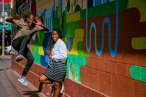 A vibrant urban scene featuring two people interacting with a colorful graffiti wall. One person is energetically jumping with a playful expression, while the other stands casually leaning against the wall, smiling. The mural displays a variety of abstract shapes and bold colors.