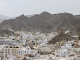 A cityscape with a mix of white and beige buildings filling the foreground, nestled at the base of a rugged mountain range. The architecture features flat roofs and mosque domes, characteristic of Middle Eastern design. The mountains in the background are steep and rocky, creating a stark contrast with the urban environment.