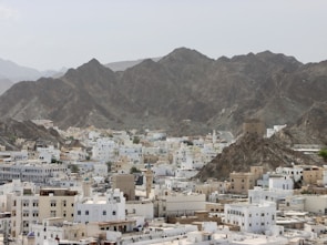 A cityscape with a mix of white and beige buildings filling the foreground, nestled at the base of a rugged mountain range. The architecture features flat roofs and mosque domes, characteristic of Middle Eastern design. The mountains in the background are steep and rocky, creating a stark contrast with the urban environment.