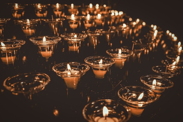 Rows of lit candles are arranged in a dark setting, each one in a transparent glass holder, casting a warm glow and soft reflections on the surrounding surfaces.