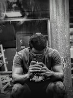 A person sitting in a gym environment, holding a smartphone with both hands and looking at it intently. The setting is dimly lit with visible gym equipment like weight racks and a large pillar in the background.
