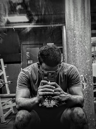 A person sitting in a gym environment, holding a smartphone with both hands and looking at it intently. The setting is dimly lit with visible gym equipment like weight racks and a large pillar in the background.