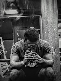 A person sitting in a gym environment, holding a smartphone with both hands and looking at it intently. The setting is dimly lit with visible gym equipment like weight racks and a large pillar in the background.