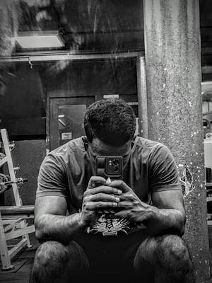 A person sitting in a gym environment, holding a smartphone with both hands and looking at it intently. The setting is dimly lit with visible gym equipment like weight racks and a large pillar in the background.