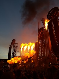 A concert stage with large flame effects shooting into the air. The setting appears to be outdoors during a performance, with an elaborate stage setup and a crowd of people. The sky is dusky, transitioning from day to night, creating a dramatic backdrop.