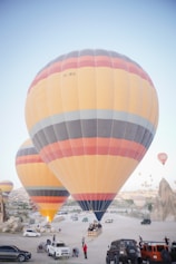 Multiple large hot air balloons are prepared for flight in a desert-like landscape with rocky formations. Several vehicles, including cars and jeeps, are parked around the area, with people gathered near the balloons. The scene conveys a sense of adventure and exploration.
