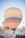 Multiple large hot air balloons are prepared for flight in a desert-like landscape with rocky formations. Several vehicles, including cars and jeeps, are parked around the area, with people gathered near the balloons. The scene conveys a sense of adventure and exploration.
