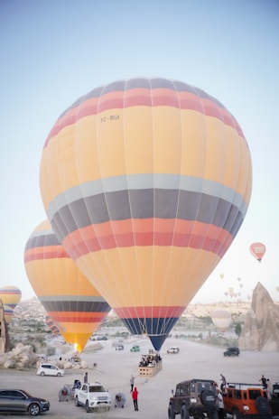 Multiple large hot air balloons are prepared for flight in a desert-like landscape with rocky formations. Several vehicles, including cars and jeeps, are parked around the area, with people gathered near the balloons. The scene conveys a sense of adventure and exploration.