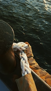 A thick, braided rope is coiled around a metallic cleat on a boat. The rope rests on a surface covered by a protective brown tarp, with the dark blue water in the background reflecting the light.