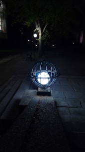 A glowing spherical light fixture enclosed in a metal cage on the ground at night, illuminating the surrounding area. Nearby is a tree, and other similar lights are seen in the background, casting dim glows in the darkness.