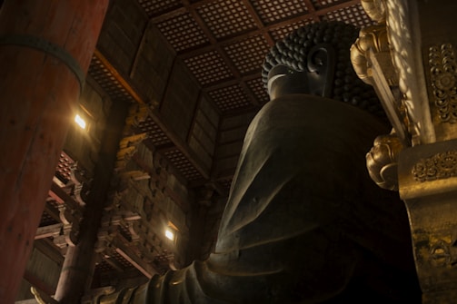 A large statue of Buddha occupies the right side of the frame, with intricate details visible on the golden robes. The statue is housed within a spacious wooden structure featuring an ornate ceiling and large supporting pillars. Light filters in from small windows, highlighting the textures and architectural elements.