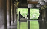 A serene interior space features large wooden columns, a polished floor reflecting light, and several traditional lanterns hanging from the ceiling. A silhouette of a person stands in front of a Buddha statue, set against a backdrop of lush green foliage visible through open architectural elements.