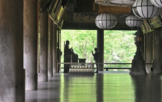 A serene interior space features large wooden columns, a polished floor reflecting light, and several traditional lanterns hanging from the ceiling. A silhouette of a person stands in front of a Buddha statue, set against a backdrop of lush green foliage visible through open architectural elements.