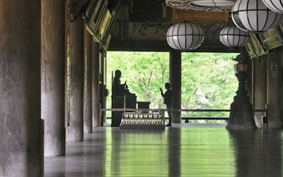 A serene interior space features large wooden columns, a polished floor reflecting light, and several traditional lanterns hanging from the ceiling. A silhouette of a person stands in front of a Buddha statue, set against a backdrop of lush green foliage visible through open architectural elements.