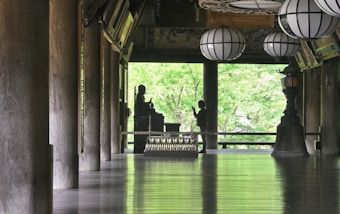 A serene interior space features large wooden columns, a polished floor reflecting light, and several traditional lanterns hanging from the ceiling. A silhouette of a person stands in front of a Buddha statue, set against a backdrop of lush green foliage visible through open architectural elements.