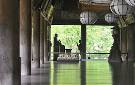 A serene interior space features large wooden columns, a polished floor reflecting light, and several traditional lanterns hanging from the ceiling. A silhouette of a person stands in front of a Buddha statue, set against a backdrop of lush green foliage visible through open architectural elements.