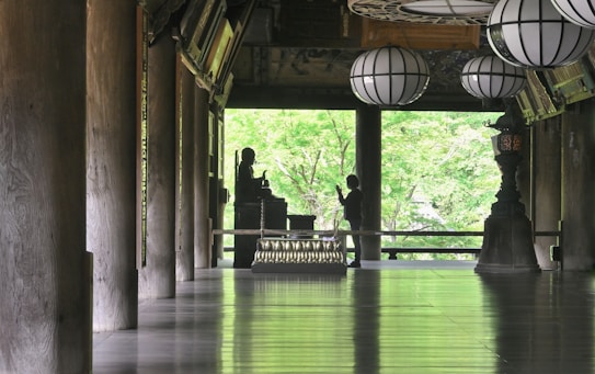 A serene interior space features large wooden columns, a polished floor reflecting light, and several traditional lanterns hanging from the ceiling. A silhouette of a person stands in front of a Buddha statue, set against a backdrop of lush green foliage visible through open architectural elements.