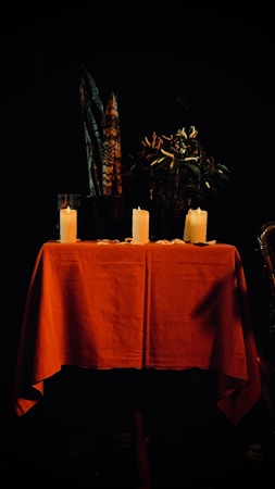 A table covered with a red cloth is the focal point, featuring three candles standing upright. Behind the table, there is a plant with dark green leaves, and further back, tall decorative feathers are positioned. The setting is dimly lit, creating a moody and intimate ambiance.