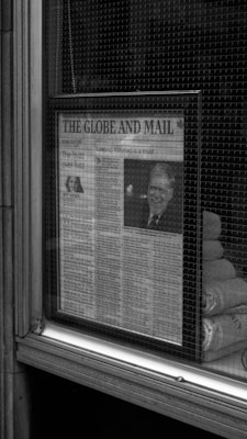 A newspaper, titled The Globe and Mail, is displayed behind a glass window. The main article features a black and white image of a person. Reflective surfaces and the texture of the window mesh are visible, suggesting it is affixed to a building's exterior.