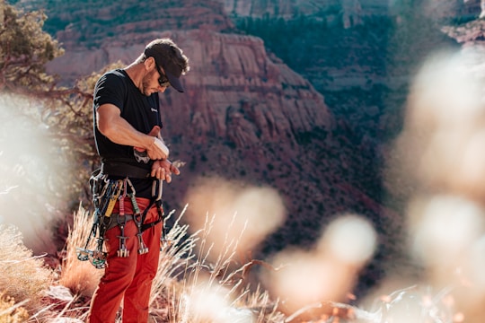 A person is preparing for rock climbing, adjusting their gear with a scenic backdrop of rugged canyon landscape. The climber is dressed in a black shirt, red pants, and is surrounded by climbing equipment such as carabiners and ropes. The environment is dry with some vegetation in the foreground, mostly yellowish grass and trees.