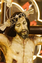 A detailed sculpture of a crucifixion featuring a figure with a crown of thorns and a pained expression. The sculpture is realistically painted, showing wounds and blood. The background includes illuminated designs, creating a dramatic contrast with the figure.