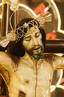 A detailed sculpture of a crucifixion featuring a figure with a crown of thorns and a pained expression. The sculpture is realistically painted, showing wounds and blood. The background includes illuminated designs, creating a dramatic contrast with the figure.