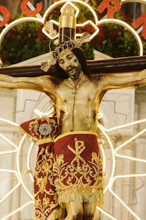 A religious statue depicts Jesus on the cross, adorned with an intricate golden crown and wearing a richly decorated red cloth with gold embroidery. The background features illuminated circular patterns and hints of greenery and flowers.